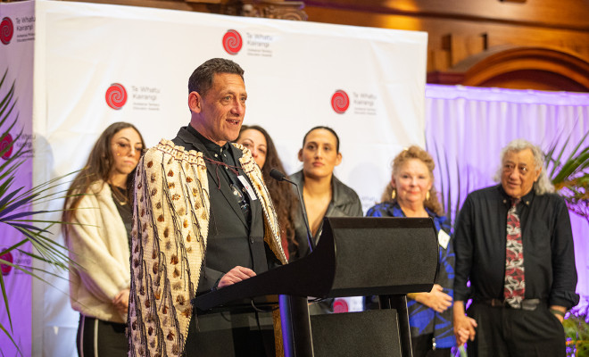 Dr Ēnoka Murphy standing in front of a podium, wearing the korowai Rauaroha that is awarded annually to the Prime Minister’s Educator of the Year 