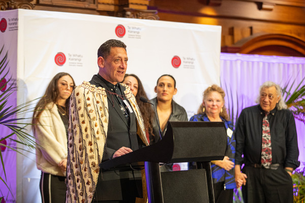 Dr Ēnoka Murphy standing in front of a podium, wearing the korowai Rauaroha that is awarded annually to the Prime Minister’s Educator of the Year 