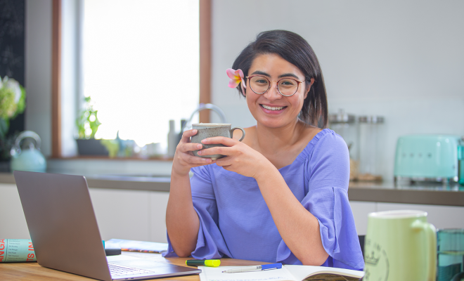 Woman sitting in front of a laptop and notebook smiling and holding a mug.