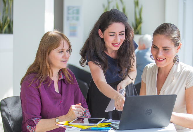 A woman points to a laptop screen, showing two other women assistive technology resources on the screen.